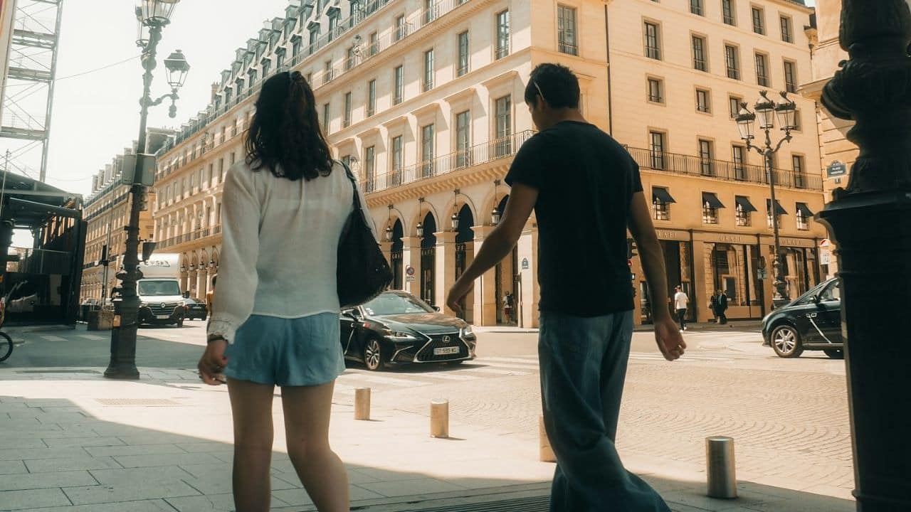Two people walk away from the camera down a sunny city street with tall buildings.