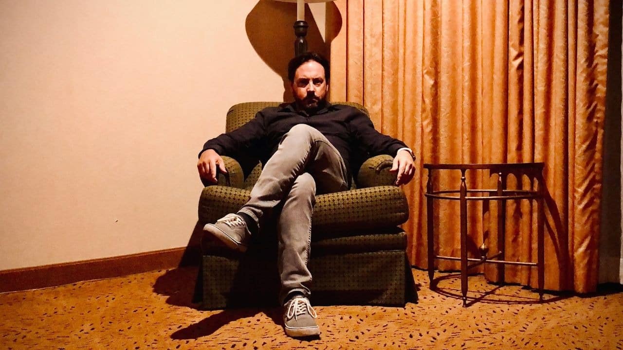 A man with a dark beard sits with crossed legs in a green patterned armchair.