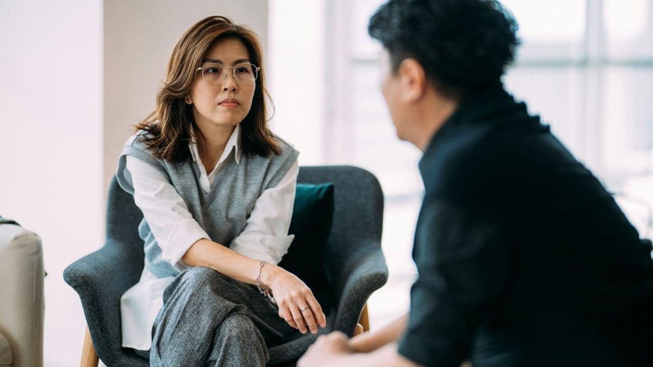 A woman in glasses listens with a neutral expression while a man speaks to her.