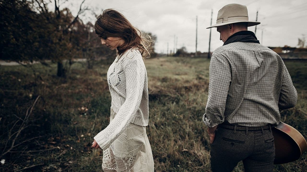 A woman in white and a man in a hat walk through a grassy field.