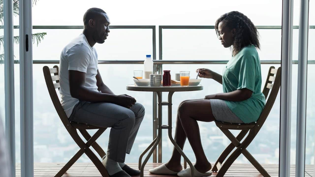 A man and woman sit at a small outdoor table eating breakfast in silence.