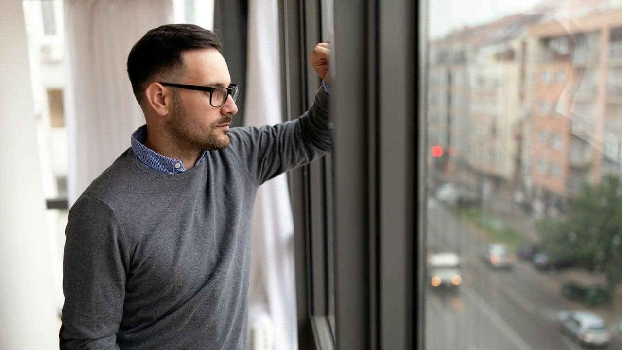 A man wearing glasses looks out of a window toward a city street during daytime.