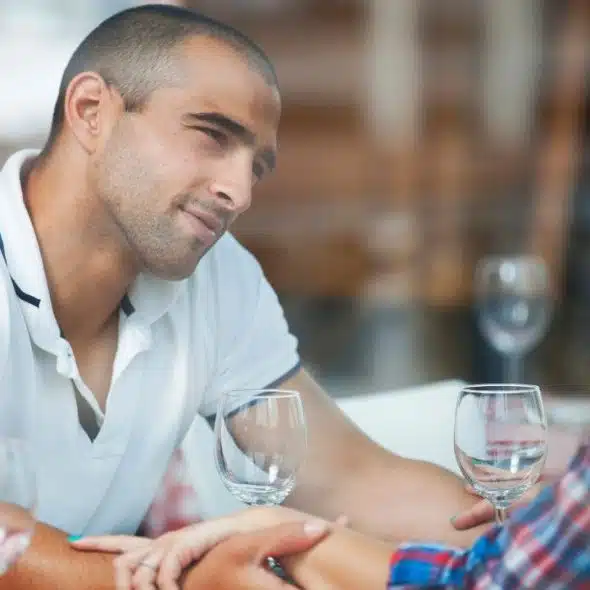 A man in a white polo shirt looks at a woman while they hold hands at dinner.