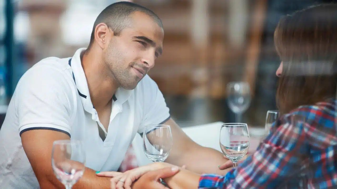 A man in a white polo shirt looks at a woman while they hold hands at dinner.