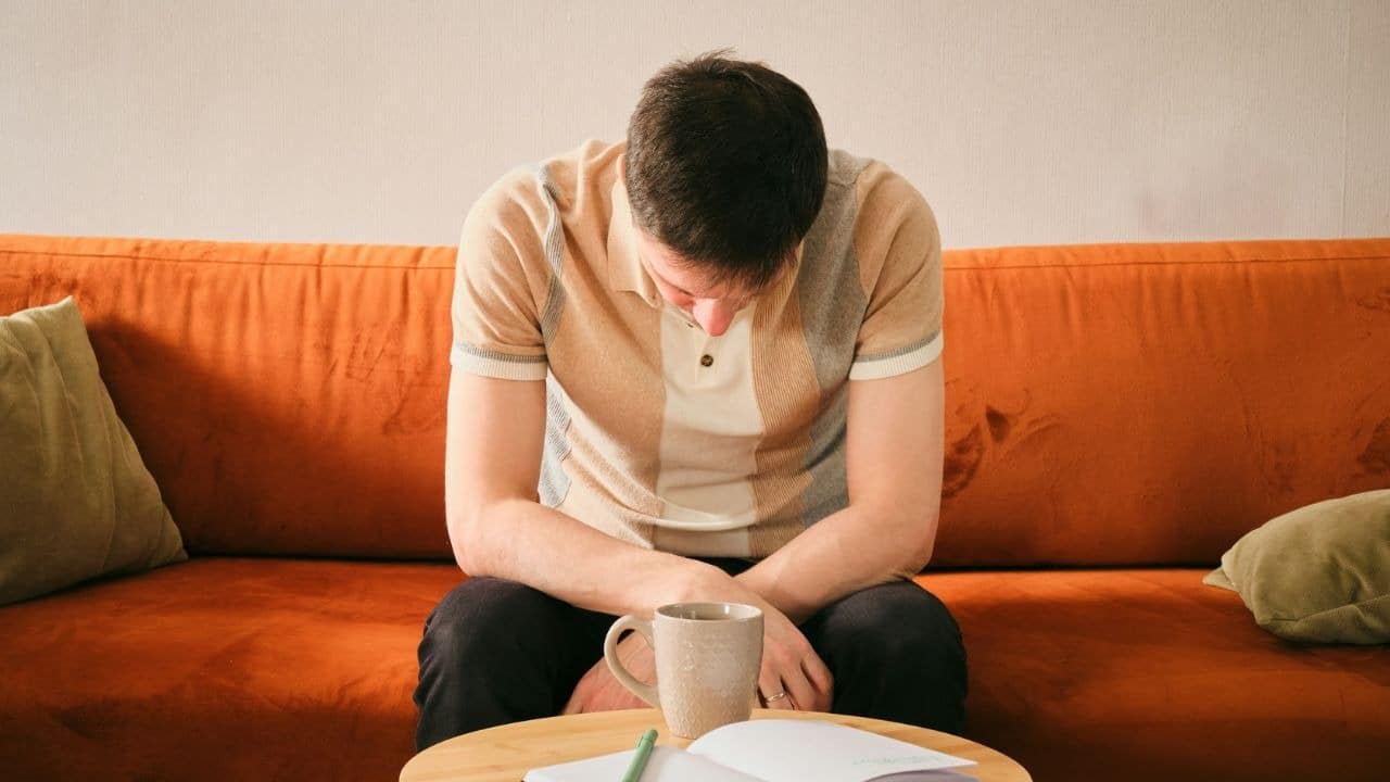 A man sits on an orange couch with his head bowed toward a mug on a table.