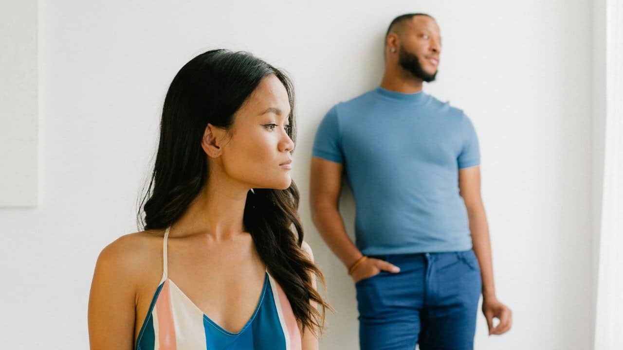 A woman looks off-camera in the foreground while a man leans against a wall in the background.