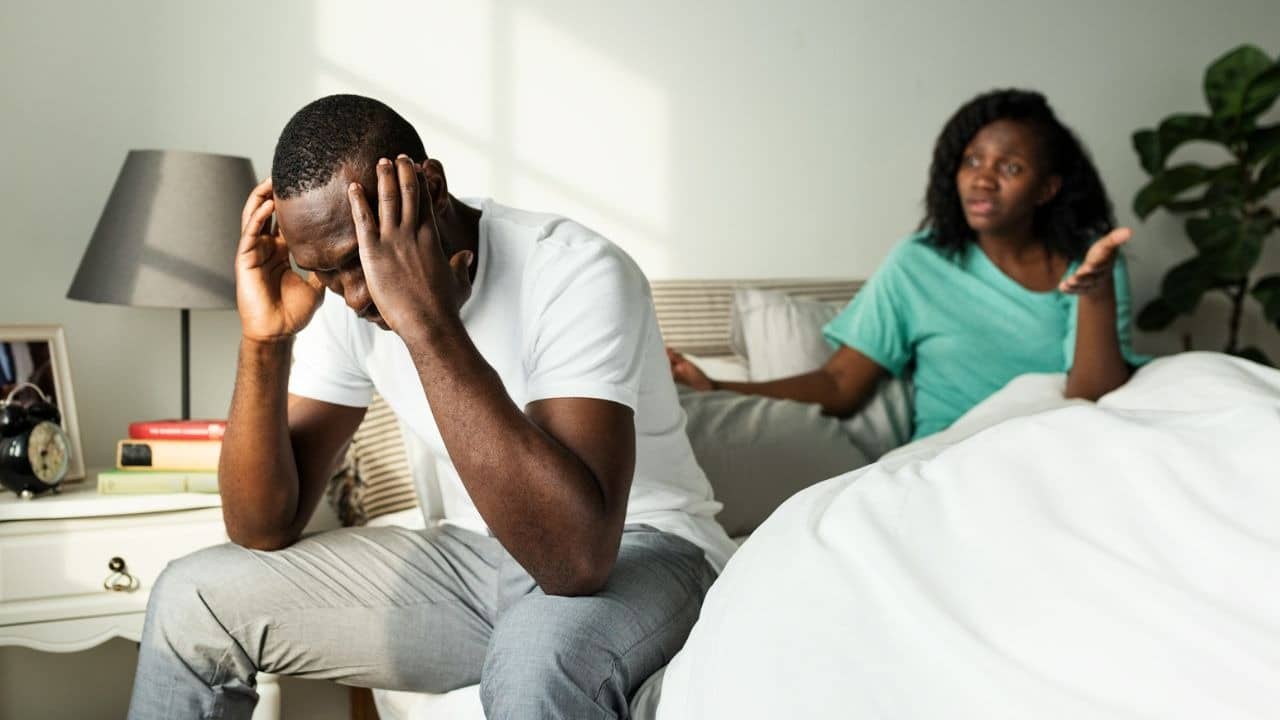 A man sits on a bed with his head in his hands while a woman speaks behind him.