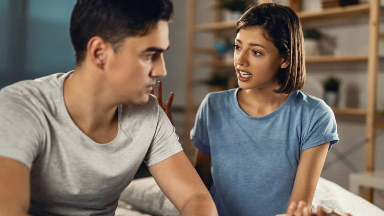 A woman in a blue shirt speaks to a man in a grey shirt indoors.
