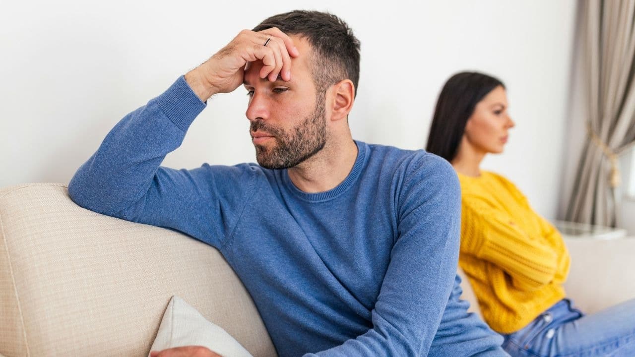 A man sits on a sofa with his hand on his forehead while a woman sits behind him.