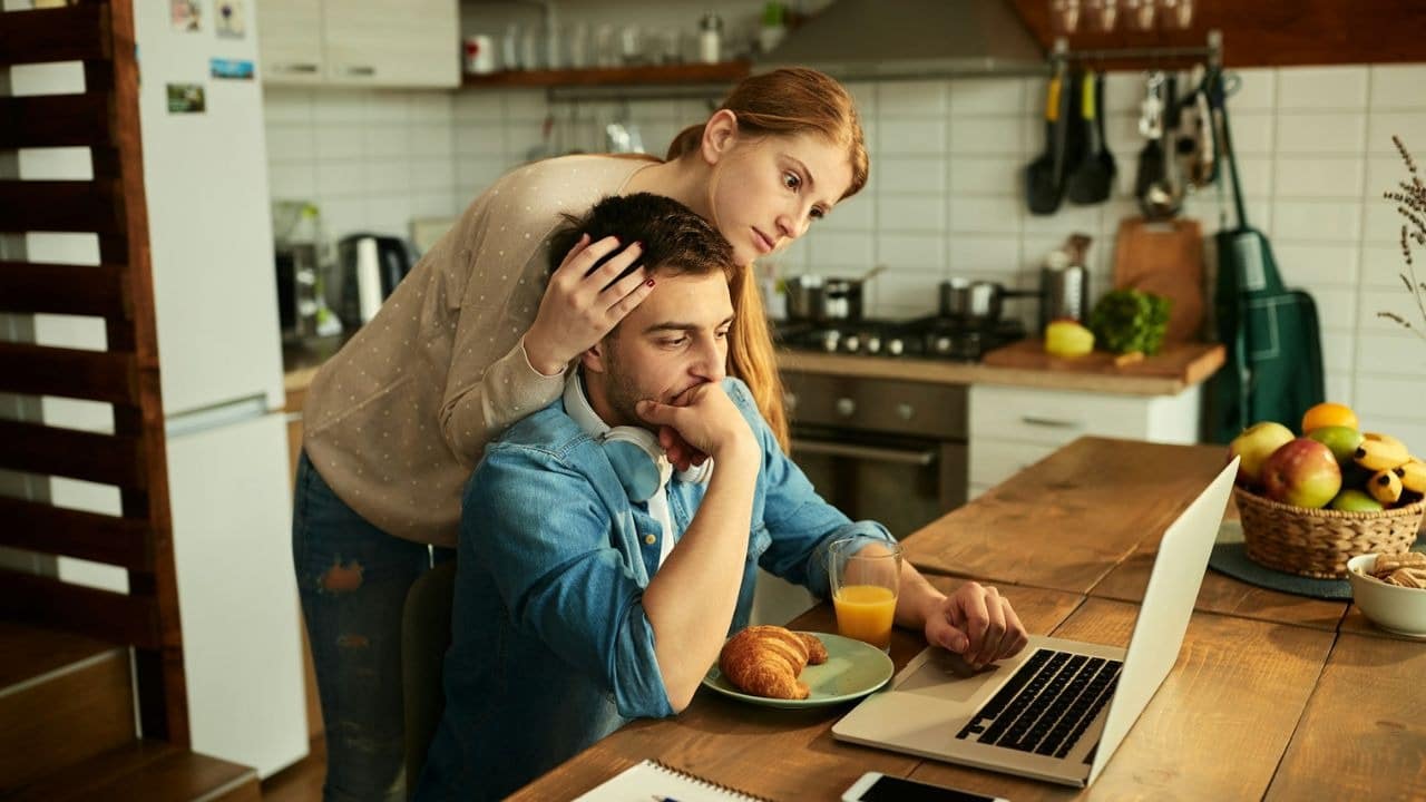 A woman leans over a man who is sitting at a kitchen table looking at a laptop.