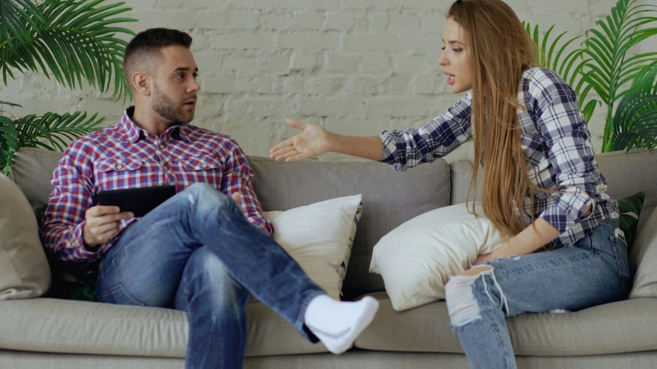 A woman gestures toward a man who is holding a tablet and looking at her on a couch.