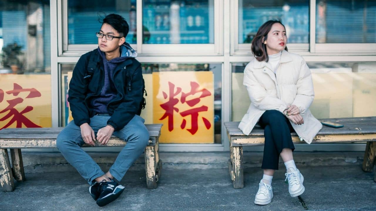 Two people sit on separate wooden benches in front of a window with yellow signs.