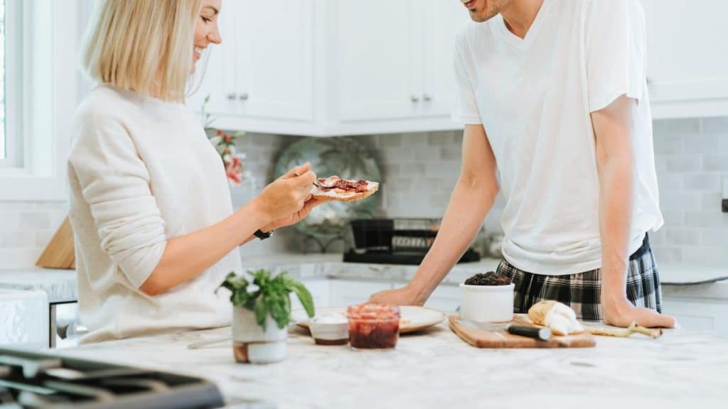 A woman spreads jam on bread while smiling at a man in the kitchen.