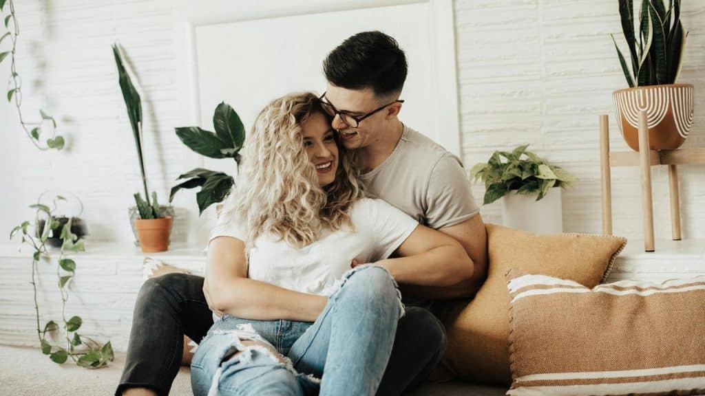 A couple sits on the floor smiling and cuddling in a cozy living room.