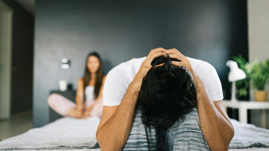 A man sits on a bed with his head in his hands while a woman sits blurred in the background.