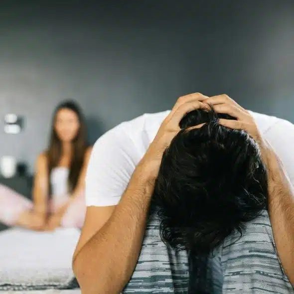 A man sits on a bed with his head in his hands while a woman sits blurred in the background.