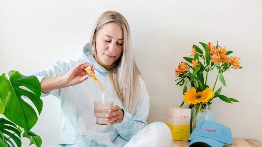 A woman in a light blue hoodie pours a drink mix into a glass.