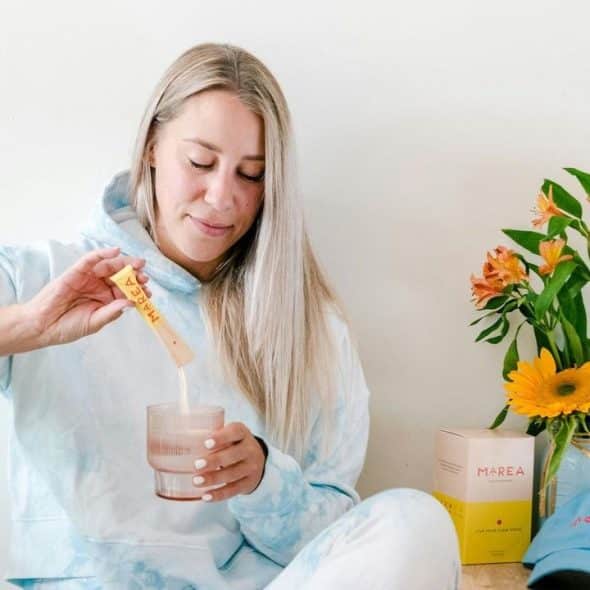 A woman in a light blue hoodie pours a drink mix into a glass.