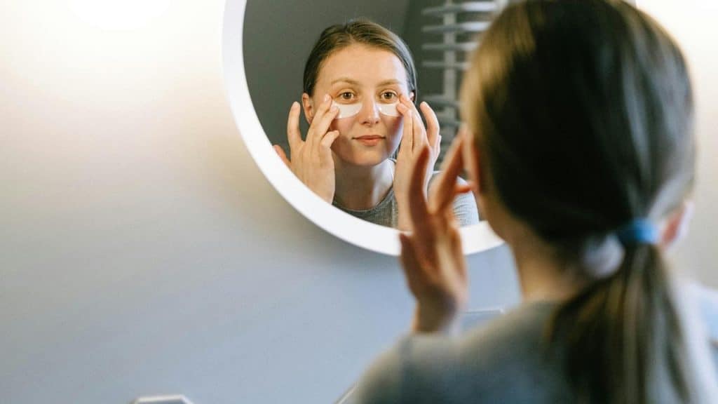 A woman applying under-eye patches while looking in a mirror.