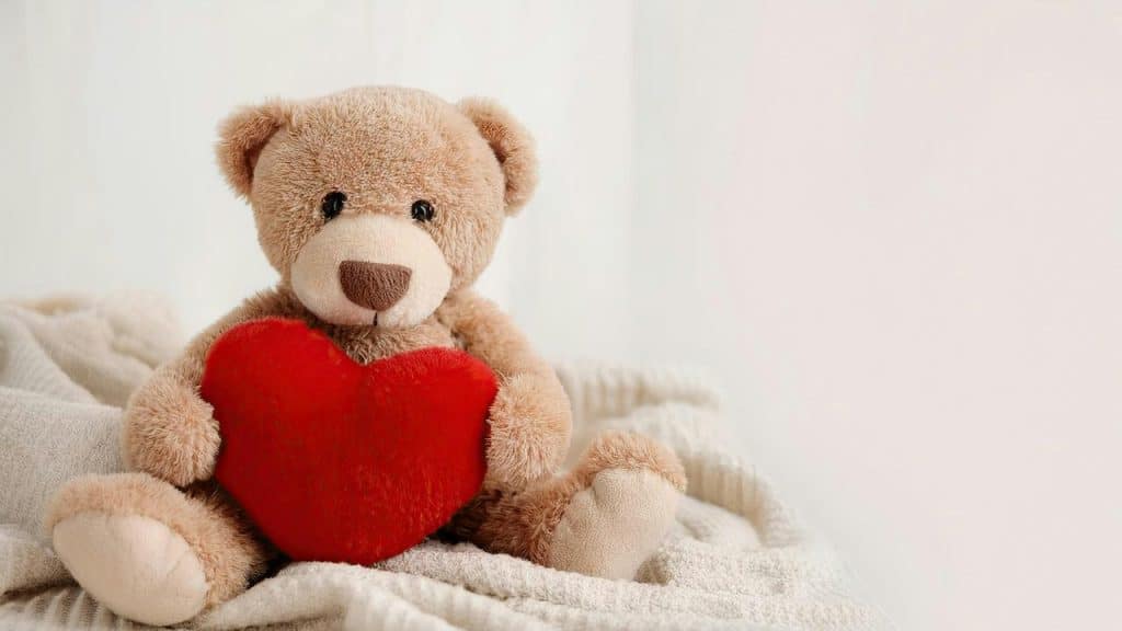 A teddy bear holds a red heart while sitting on a soft blanket.