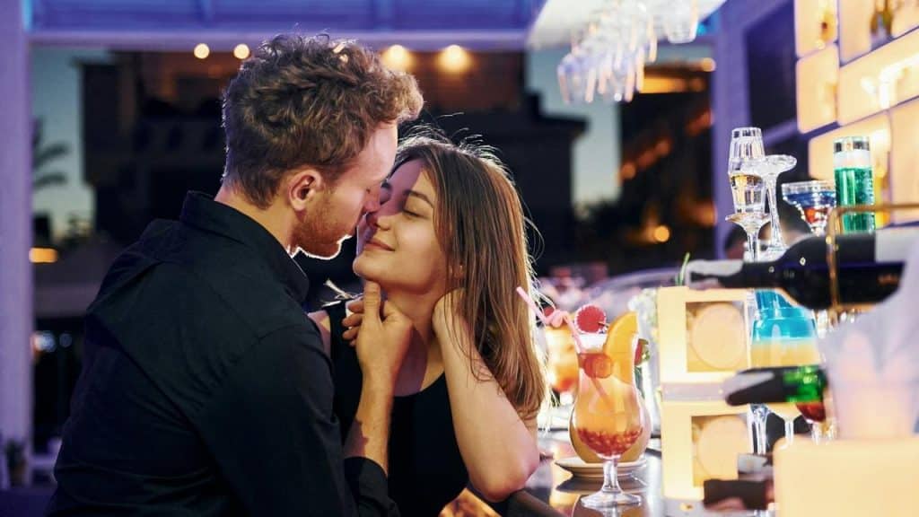 A couple kisses at a bar surrounded by colorful cocktails and lights.