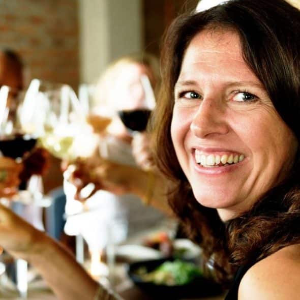 A smiling woman holding a glass of wine at a lively restaurant gathering.