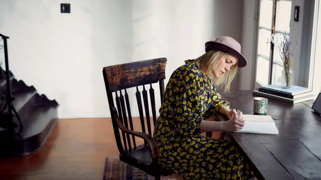 A woman wearing a hat writing in a notebook at a wooden table.