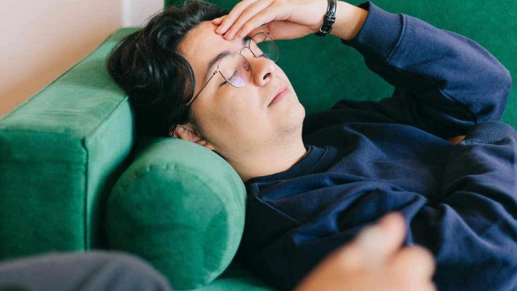 A man lies on a green couch with his hand on his forehead.