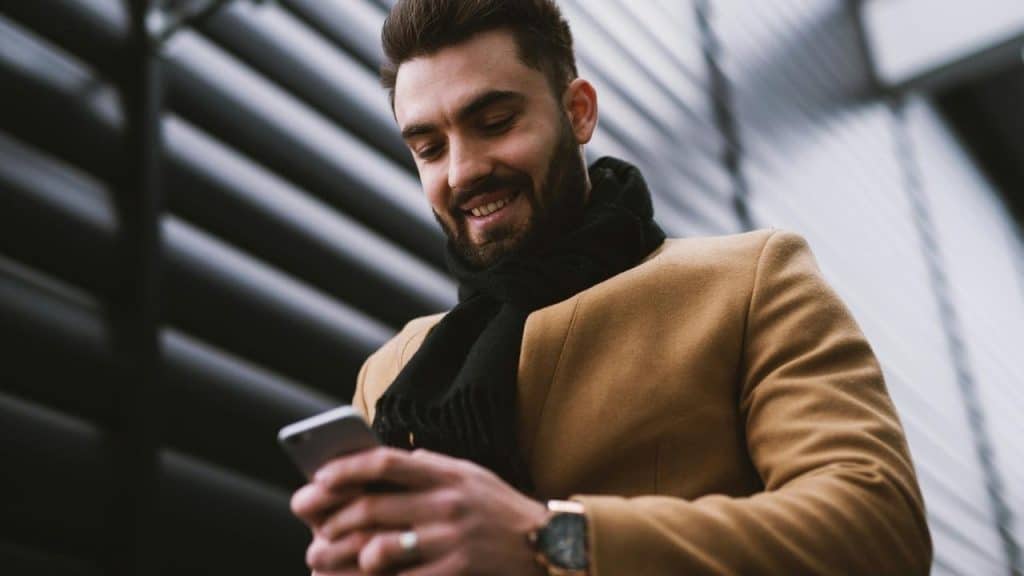 A man smiles while looking at his phone outdoors.