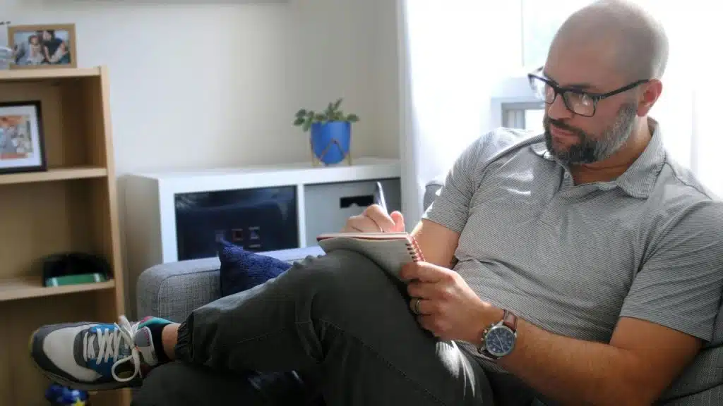 A man sits on a couch writing in a notebook