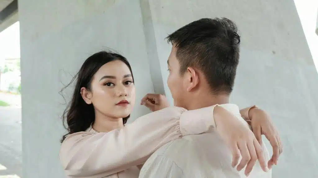 A woman looking towards the camera with her arms placed on a man’s shoulders.
