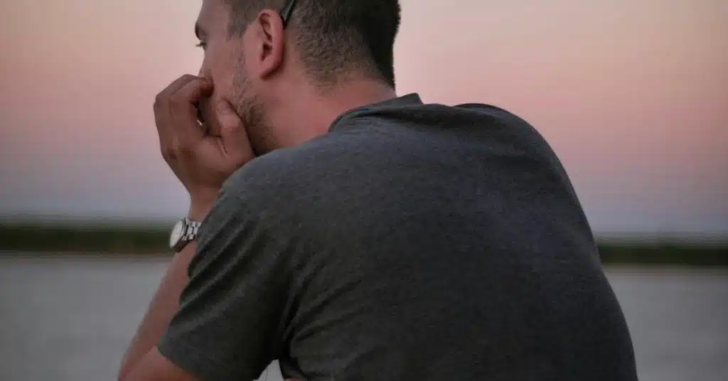 A man seen from behind, resting his chin on his hand while looking out over calm water at dusk.