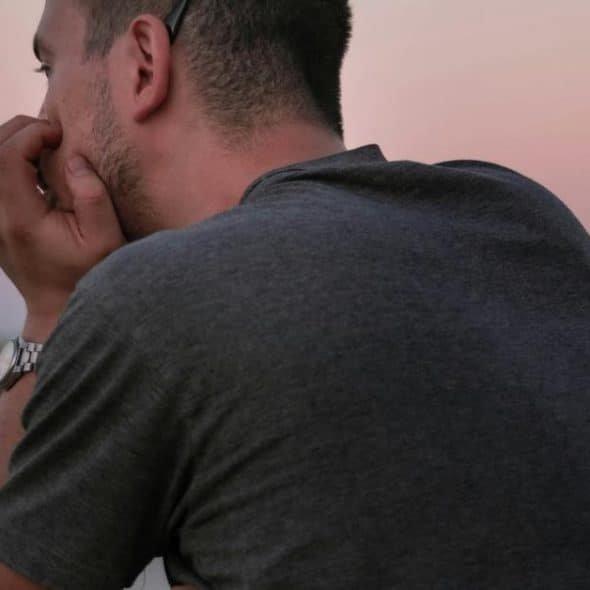 A man seen from behind, resting his chin on his hand while looking out over calm water at dusk.