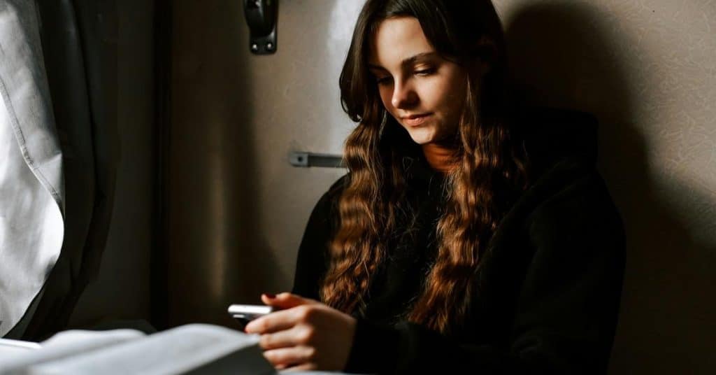 A young woman sitting by a window, looking down at her phone in soft light.
