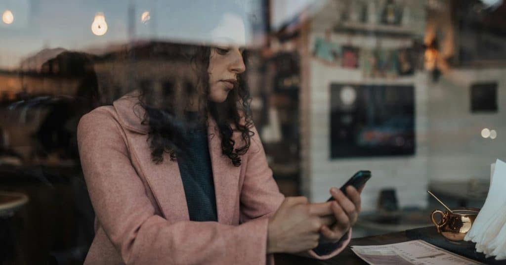 A woman sitting in a café, looking at her phone through a window reflection.