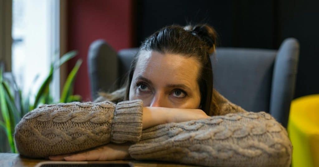 A woman resting her head on folded arms, gazing upward with a pensive expression indoors.