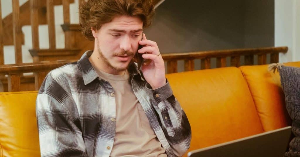 A man sitting on a couch, talking on the phone while looking at a laptop.