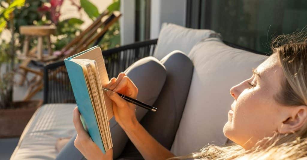 A woman reclining outdoors, writing in a notebook in warm sunlight.