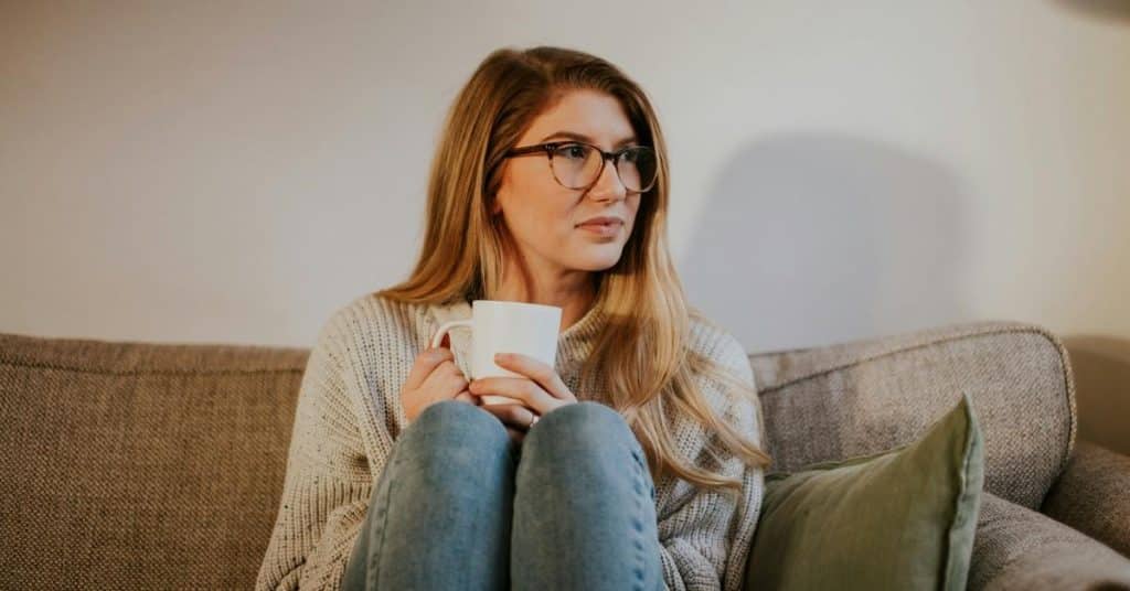 A woman wearing glasses sits on a couch holding a mug.