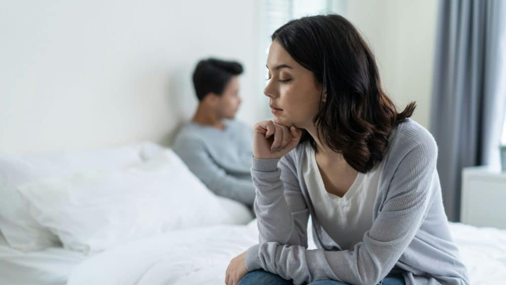 A woman is looking disappointed while sitting on the edge of a bed while her partner lies in it behind her. 