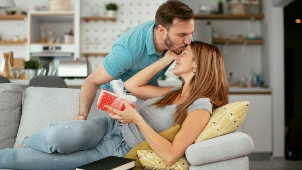 A bearded man kissing a woman’s forehead as she holds a gift and lies on a couch.