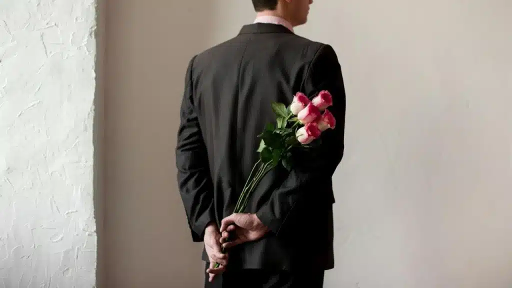 A man in a suit holding a bouquet of pink and white roses behind his back.