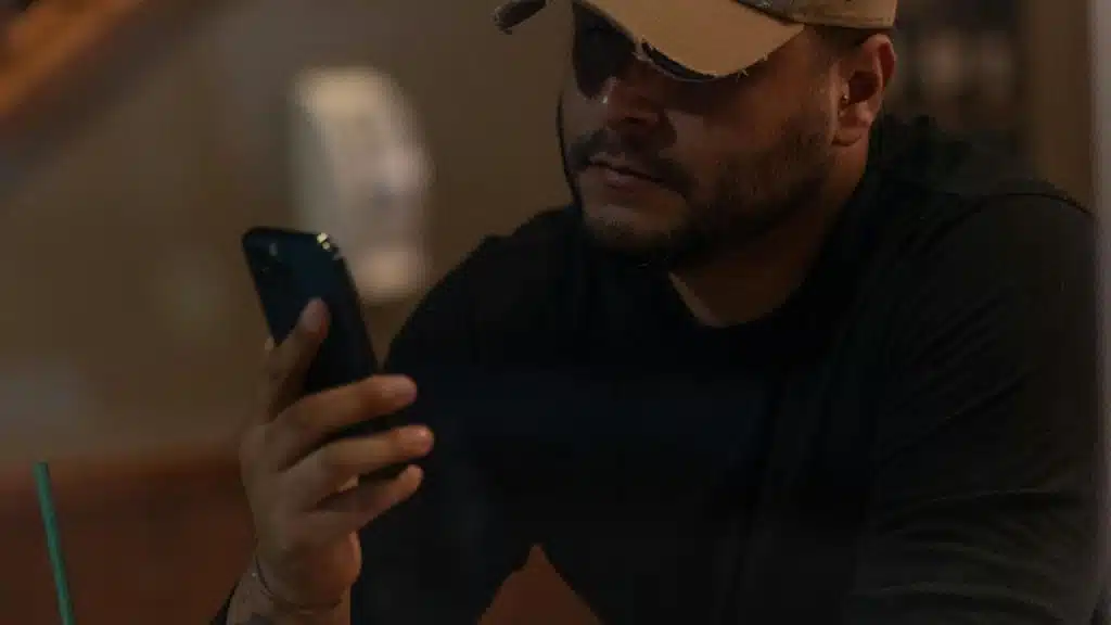 A man wearing a cap and sunglasses looks at his phone in a dimly lit café.