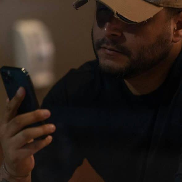 A man wearing a cap and sunglasses looks at his phone in a dimly lit café.