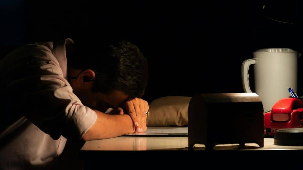 A man rests his head on his arms at a desk in a dark room.