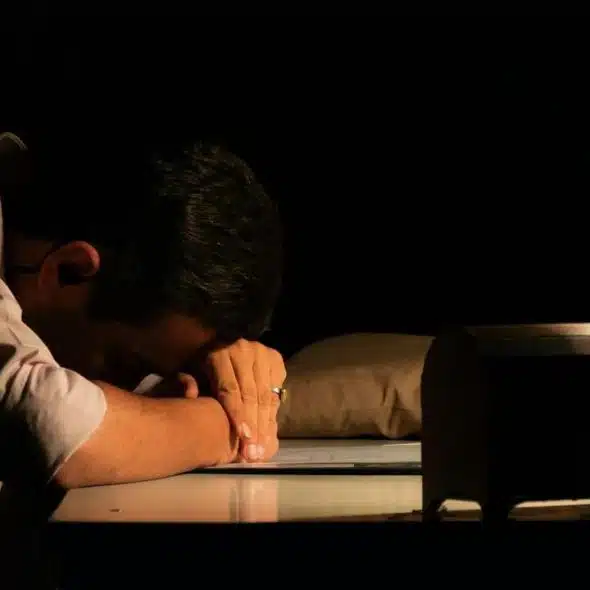A man rests his head on his arms at a desk in a dark room.