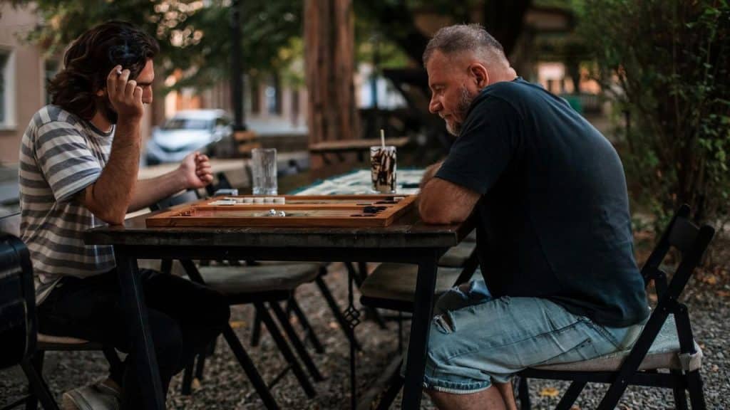 A younger man and an older man playing backgammon at an outdoor table.
