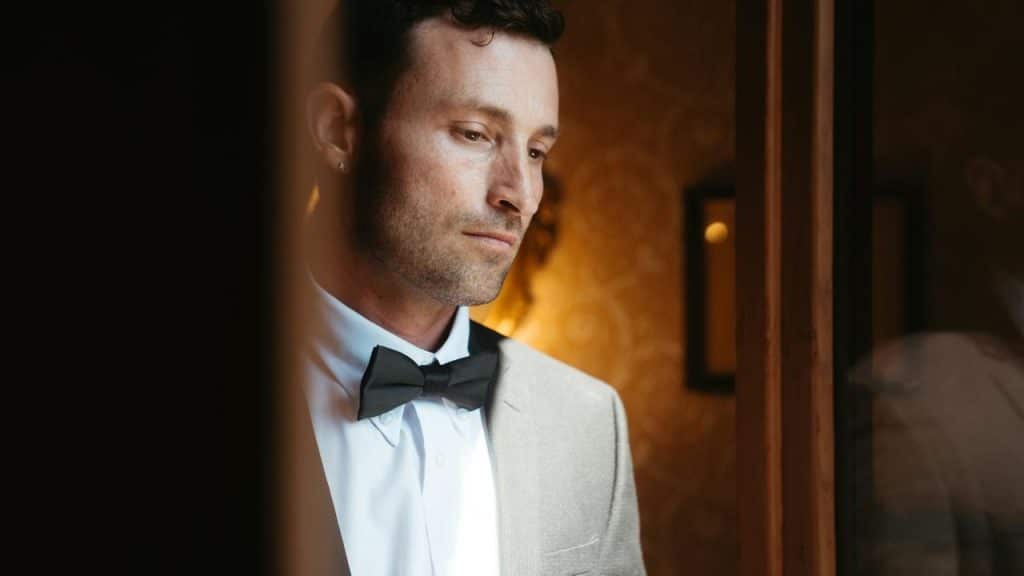 A man in a suit and bow tie looking thoughtful by a window.