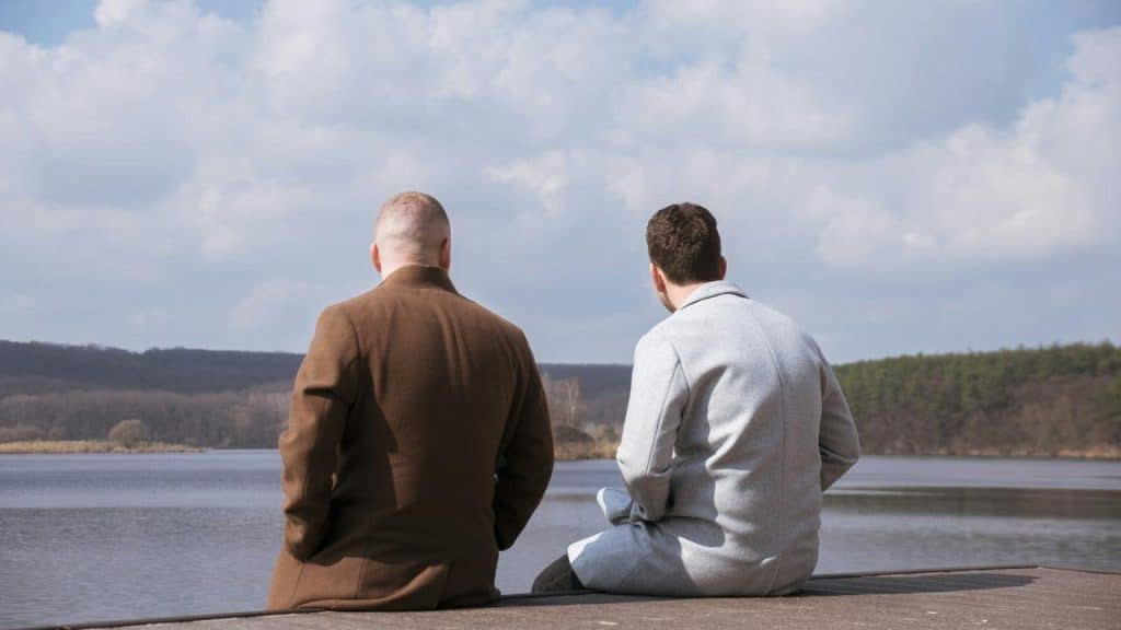 A pair of men in coats sitting on a dock facing a lake.