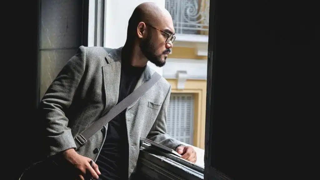 A man in glasses and a blazer leaning on a windowsill while looking outside.